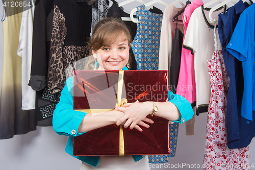 Image of Happy young girl standing with big red gift on background of clothes on hangers