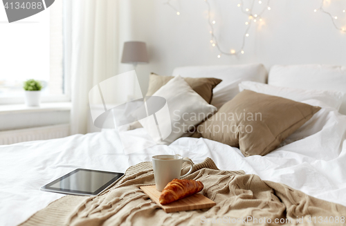 Image of tablet pc, coffee cup and croissant on bed at home