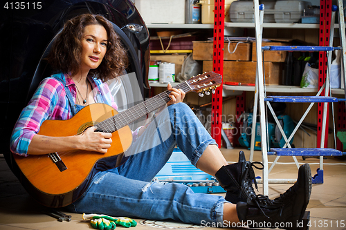 Image of beautiful Woman mechanic in blue overalls resting with a guitar