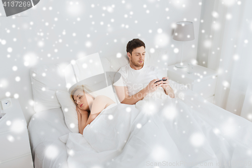 Image of man texting message while woman is sleeping in bed