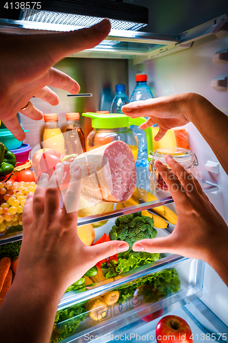 Image of Human hands reaching for food at night in the open refrigerator