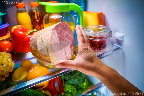 Image of Human hands reaching for food at night in the open refrigerator