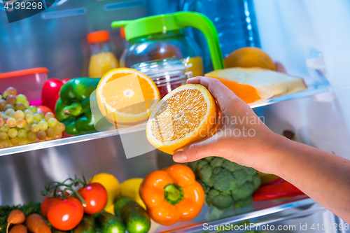 Image of Woman takes the orange from the open refrigerator.