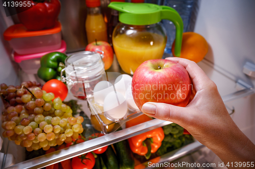 Image of Woman takes the apple from the open refrigerator.