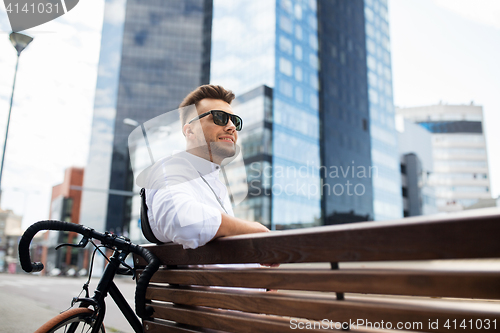 Image of happy young man with bicycle sitting on city bench