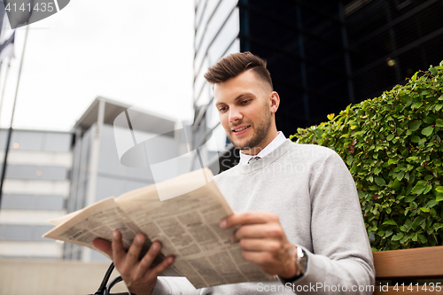 Image of smiling man reading newspaper on city street bench