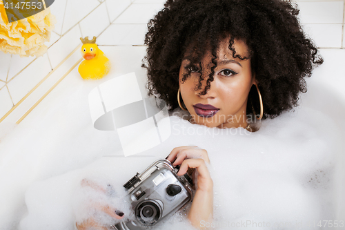 Image of young afro-american teen girl laying in bath with foam, wearing swag jewelry flawless, making selfie