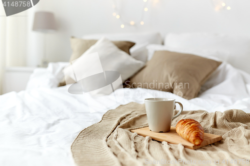Image of coffee cup and croissant on plaid in bed at home