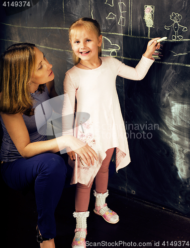 Image of portrait of mature woman teacher with little cute blonde girl pupil writing on blackboard together, lifestyle people concept