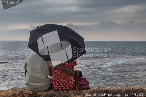 Image of Rear View of Indian couple sitting by sea with an umbrella looking at horizon.