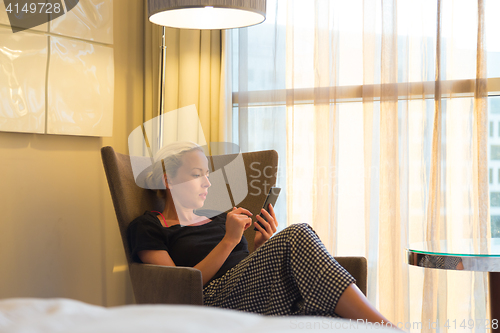 Image of Attractive woman holding and using smart phone while sitting on armchair in modern hotel room.