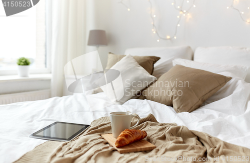 Image of tablet pc, coffee cup and croissant on bed at home
