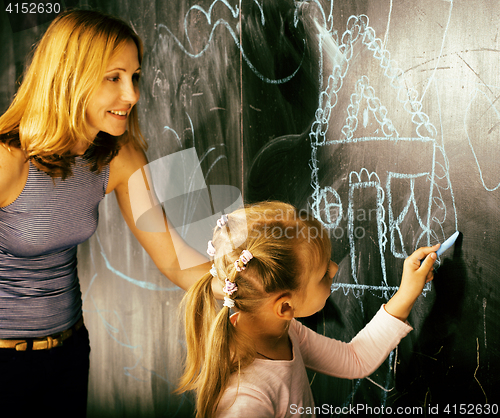 Image of portrait of mature woman teacher with little cute blonde girl pupil writing on blackboard together, lifestyle people concept