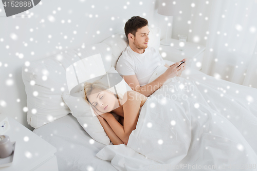 Image of man texting message while woman is sleeping in bed