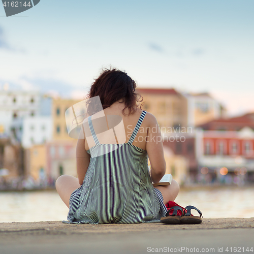 Image of Rear view of woman sitting on a pier, reading book.