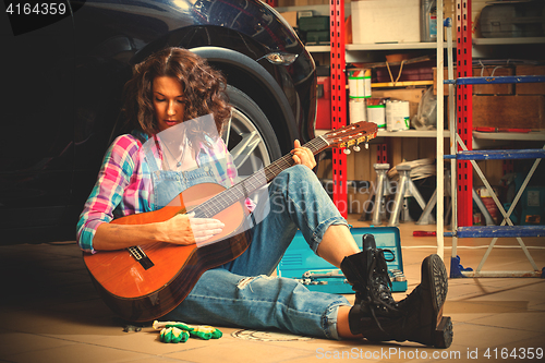 Image of beautiful brunette car mechanic with a guitar sitting in a garag