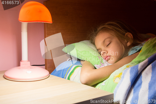 Image of Seven-year girl asleep in bed, reading lamp is included on the next table