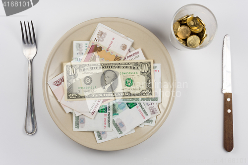 Image of On the plate is a lot of money Russian ruble and the US dollar, standing next to a glass and a fork and knife lie, top view