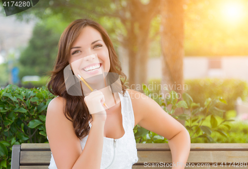 Image of Young Adult Female Student with Pencil on Bench Outdoors