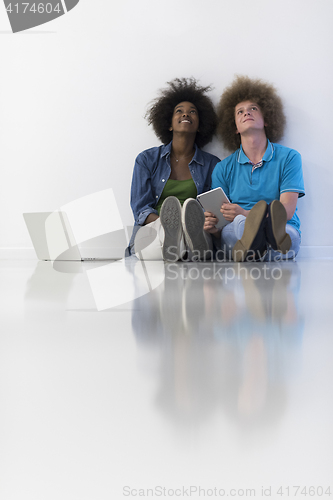Image of multiethnic couple sitting on the floor with a laptop and tablet