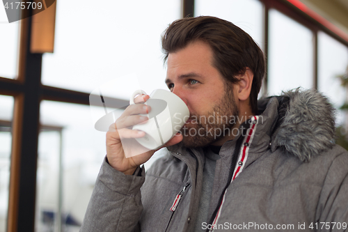 Image of Thoughtful man wearing warm winter jacket looking out the window and drinking coffee.