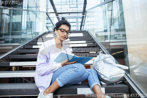 Image of young cute indian girl at university building sitting on stairs reading a book, wearing hipster glasses, lifestyle people concept