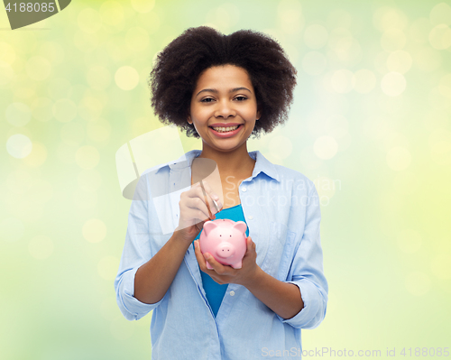 Image of happy african woman putting coin into piggy bank