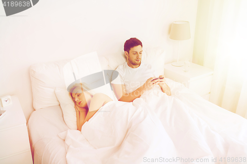 Image of man texting message while woman is sleeping in bed