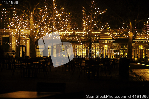 Image of empty night restaurant, lot of tables and chairs with noone, magic fairy lights on trees like christmas celebration