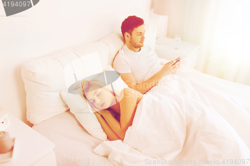 Image of man texting message while woman is sleeping in bed