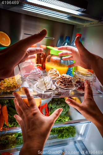 Image of Human hands reaching for sweet cake at night in the open refrige