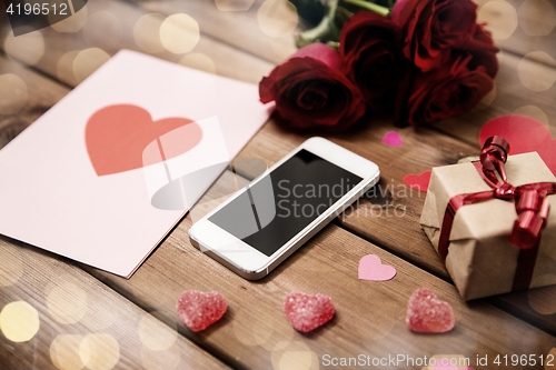 Image of close up of smartphone, gift, red roses and hearts