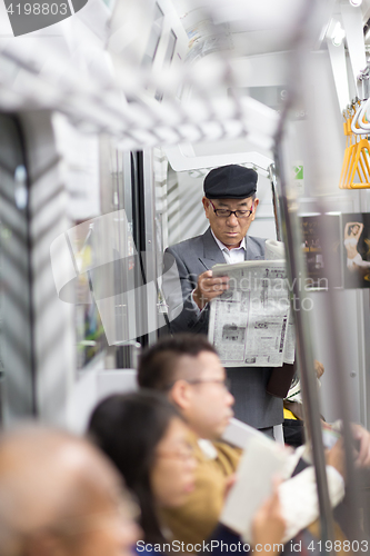 Image of Japanese businessman taking ride to work in morning, standing inside public transport and reading newspaper.