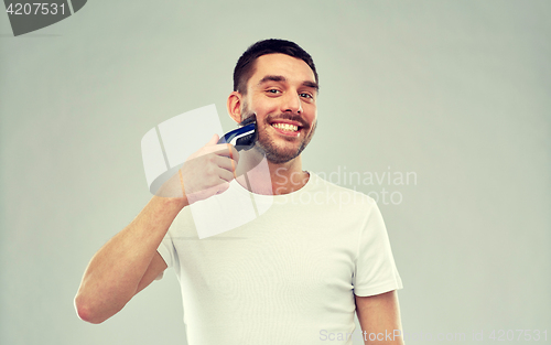 Image of smiling man shaving beard with trimmer over gray