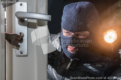 Image of Masked burglar holding flashlight while trying to break window l