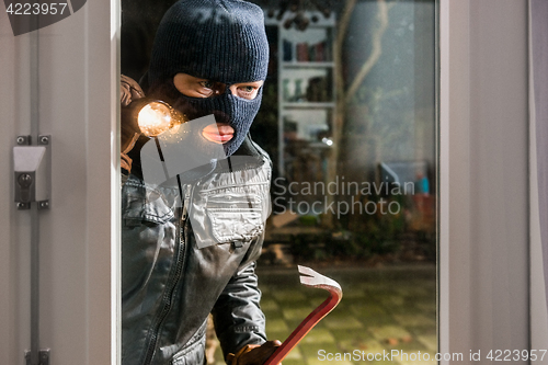 Image of Masked burglar with flashlight and crowbar looking into glass wi