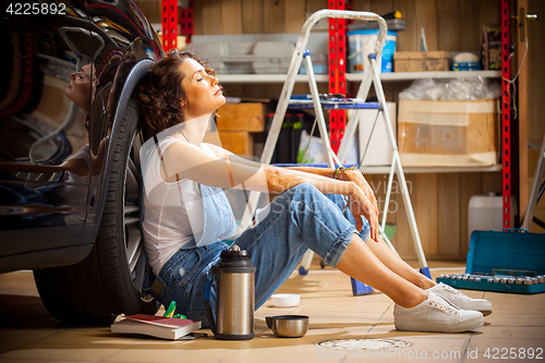 Image of woman a car mechanic sits in a blue overall in a garage near a c