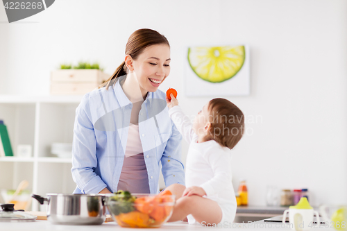 Image of baby feeding mother with carrot at home kitchen