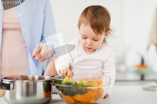 Image of baby with vegetables and mother cooking at home