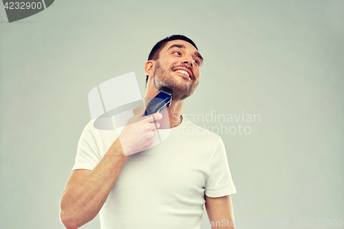Image of smiling man shaving beard with trimmer over gray