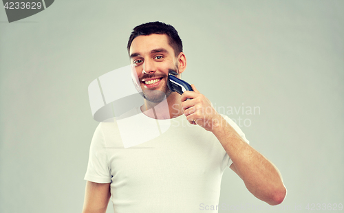 Image of smiling man shaving beard with trimmer over gray