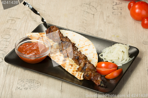 Image of shashlik. kebab skewer, black rectangular plate. sauce and onions