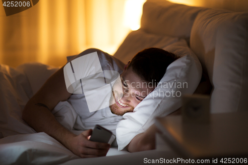 Image of happy young man with smartphone in bed at night