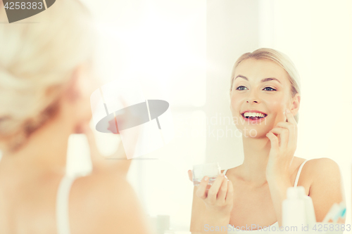 Image of happy woman applying cream to face at bathroom
