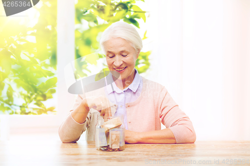 Image of senior woman putting money into glass jar at home