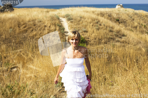 Image of portrait of beautiful blond woman in white dress standing on wind with scarf near sea