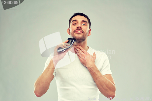 Image of smiling man shaving beard with trimmer over gray