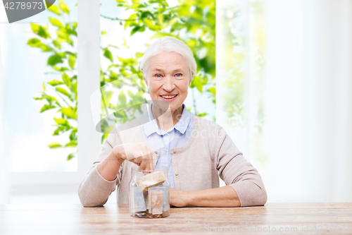 Image of senior woman putting money into glass jar at home