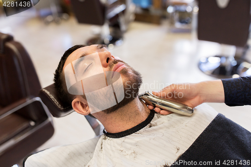 Image of man and barber with trimmer cutting beard at salon