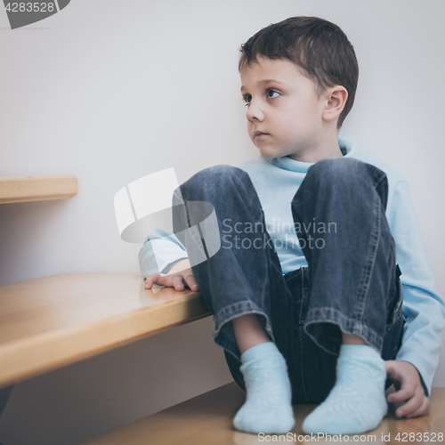 Image of one sad little boy sitting on the stairs in house at the day tim